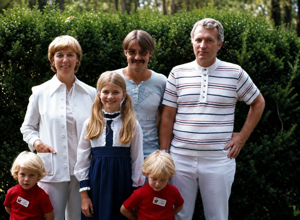 Outdoor family portrait in front of a hedge: a woman, a girl in a navy and white dress, two blond twin boys in red shirts, and two men.